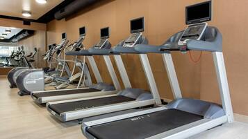 A row of treadmills with digital screens, a mirrored wall, and exercise bikes in a gym.