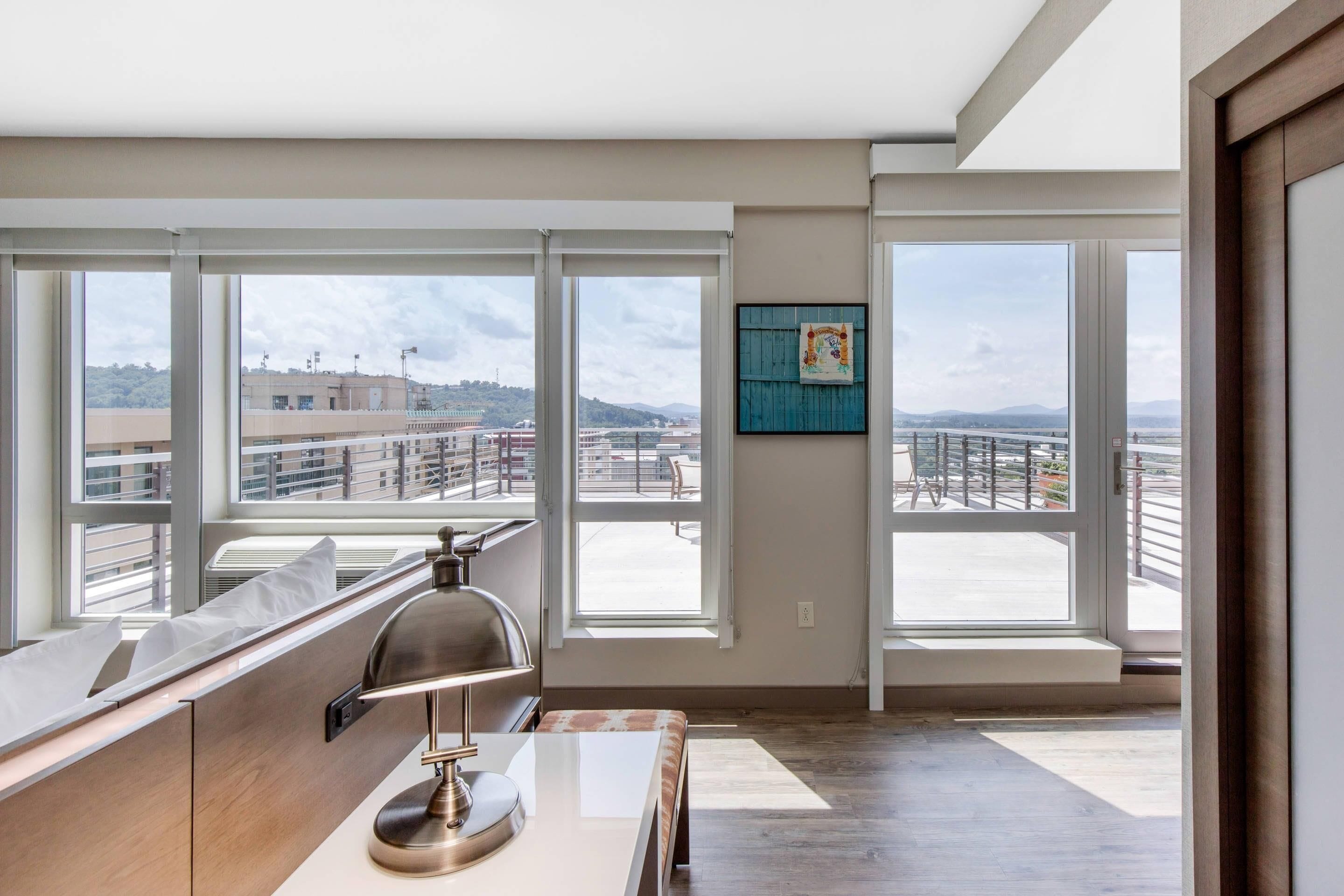 Una habitación de hotel moderna con ventanas amplias, un escritorio con lámpara y vistas a la ciudad.
