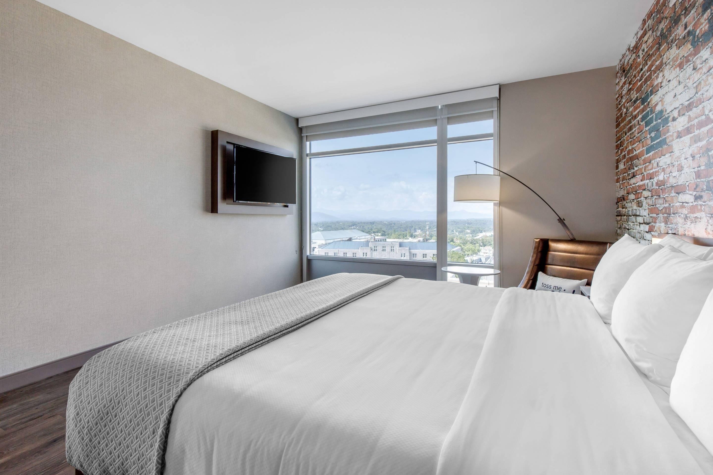 A hotel room with a large bed, a TV mounted on the wall, a window with a view of buildings, and a brick accent wall.