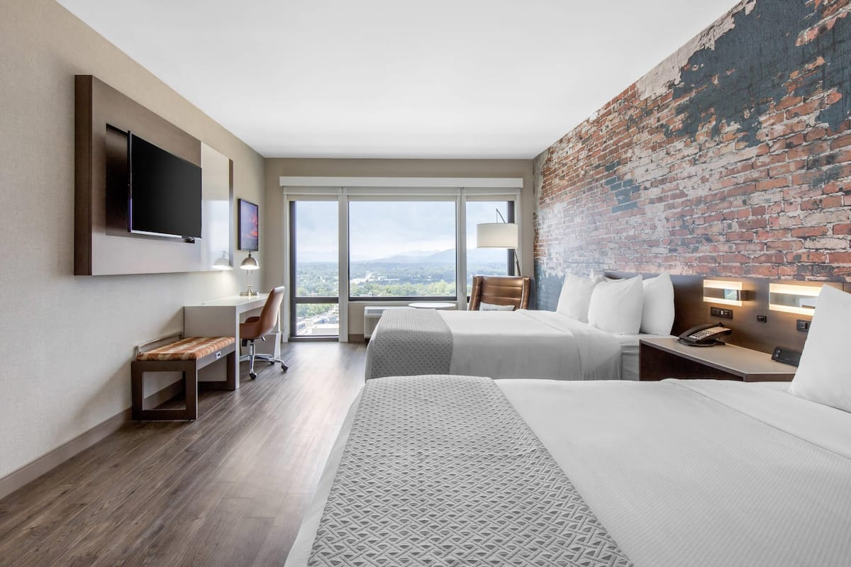 A hotel room with two beds, a desk, a TV, and a large window with a view.