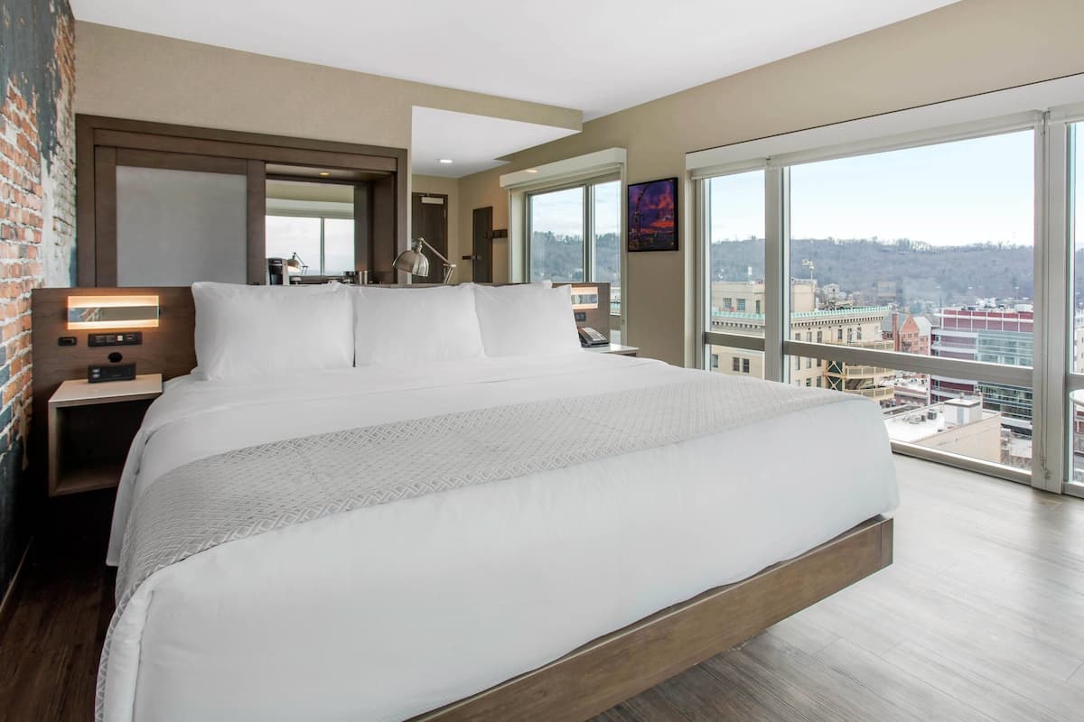 A modern hotel room with a large bed, a nightstand, a desk, and a large window offering a city view.