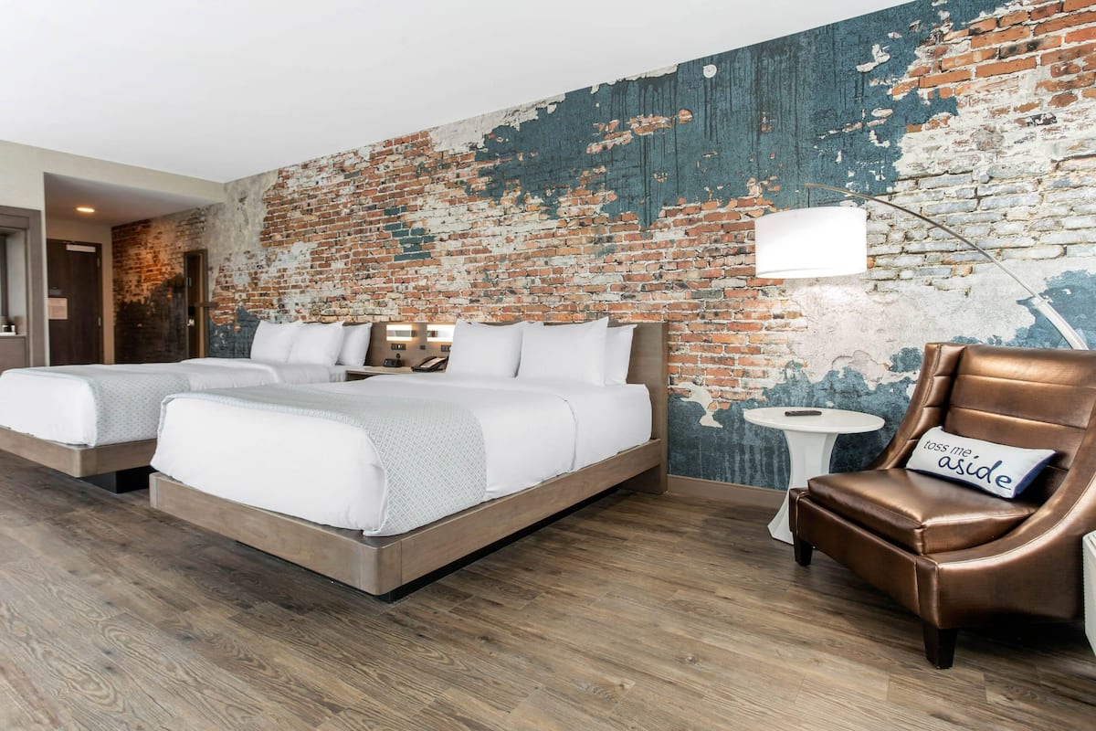 A hotel room with a brick wall, two beds, a brown leather chair, and a small round table.