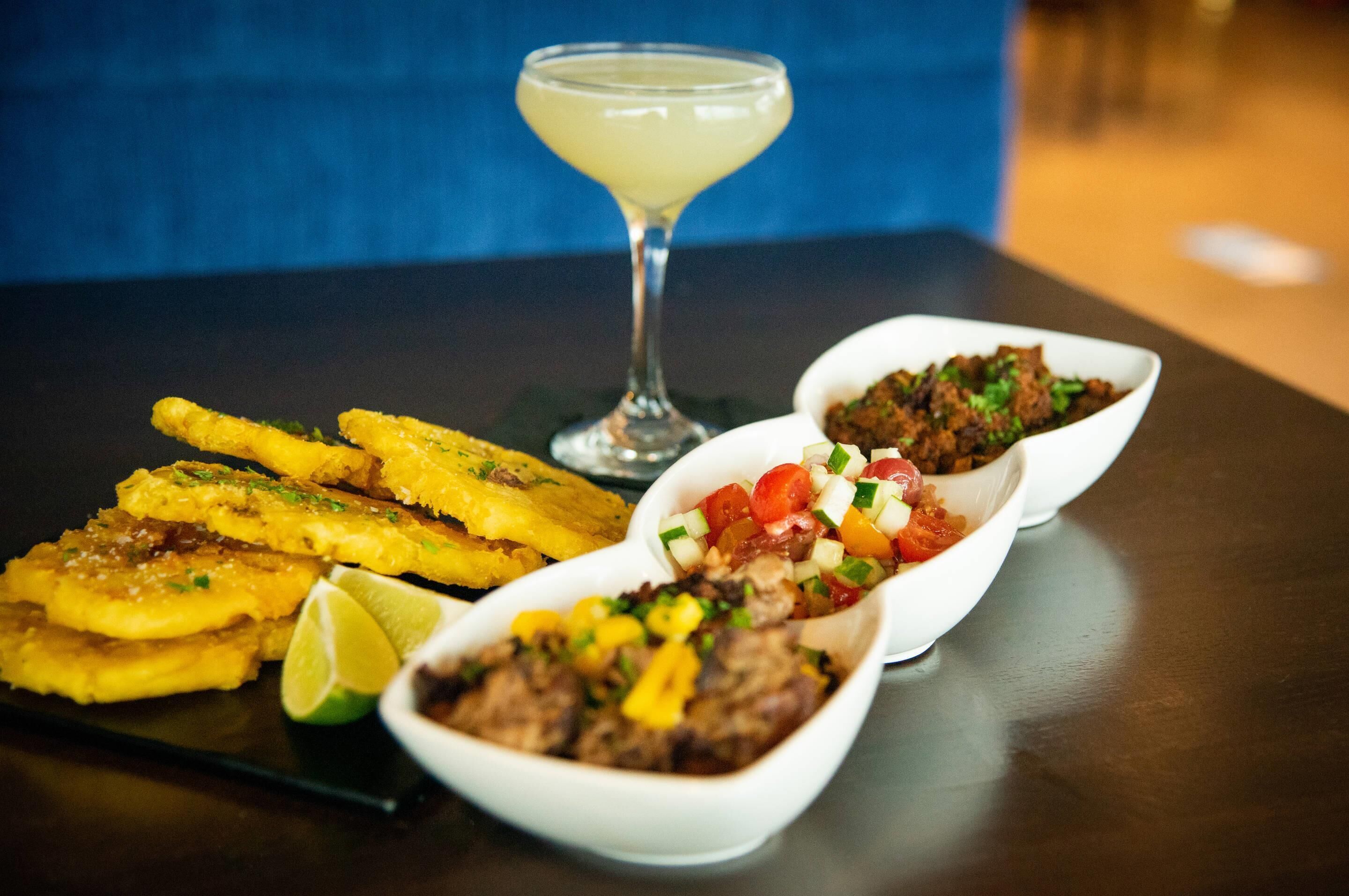 A table with three dishes: fried plantains, a meat dish with diced vegetables, and a cocktail in a martini glass.