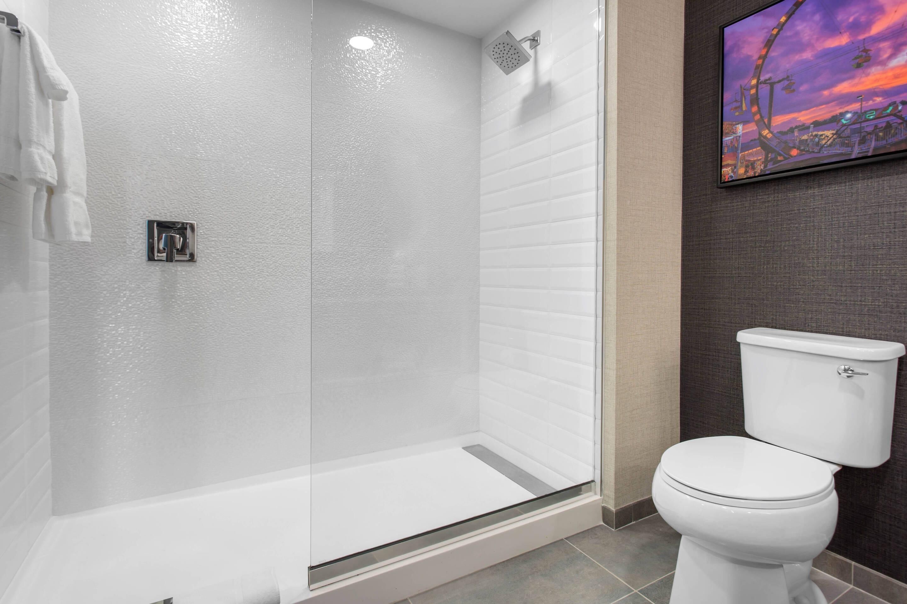 Une salle de bain dotée d’une douche en verre, de toilettes blanches et d’un téléviseur fixé au mur qui diffuse des images d’un paysage urbain.