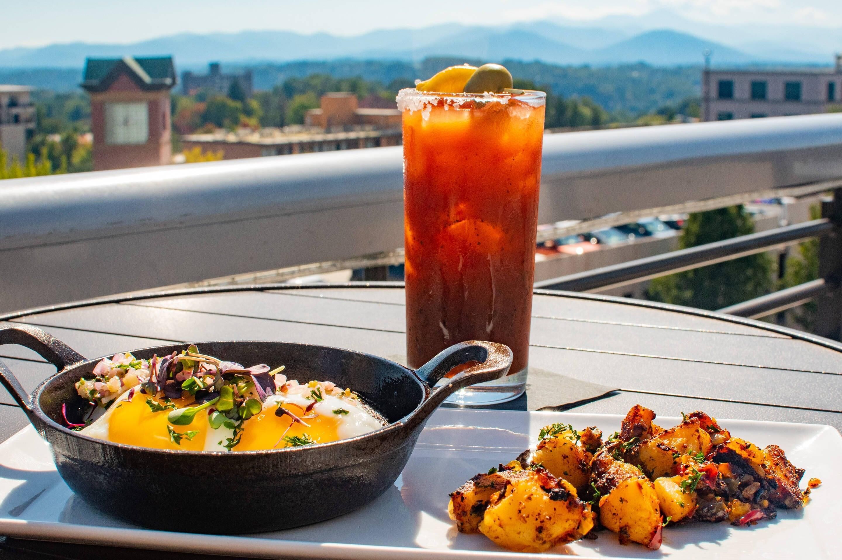 Un plato con huevos, papas asadas y un Bloody Mary, sobre una mesa con vistas panorámicas.