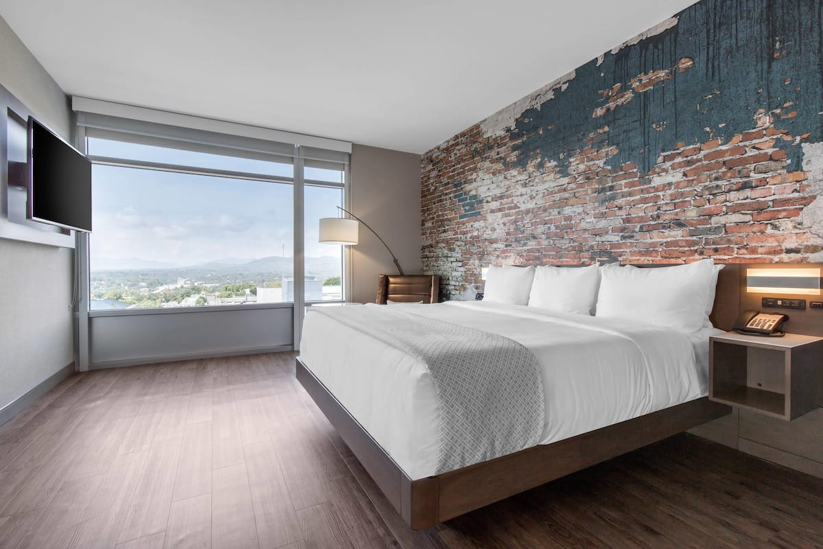 A modern bedroom with a large bed, a brick wall, a TV, and a view of the city.