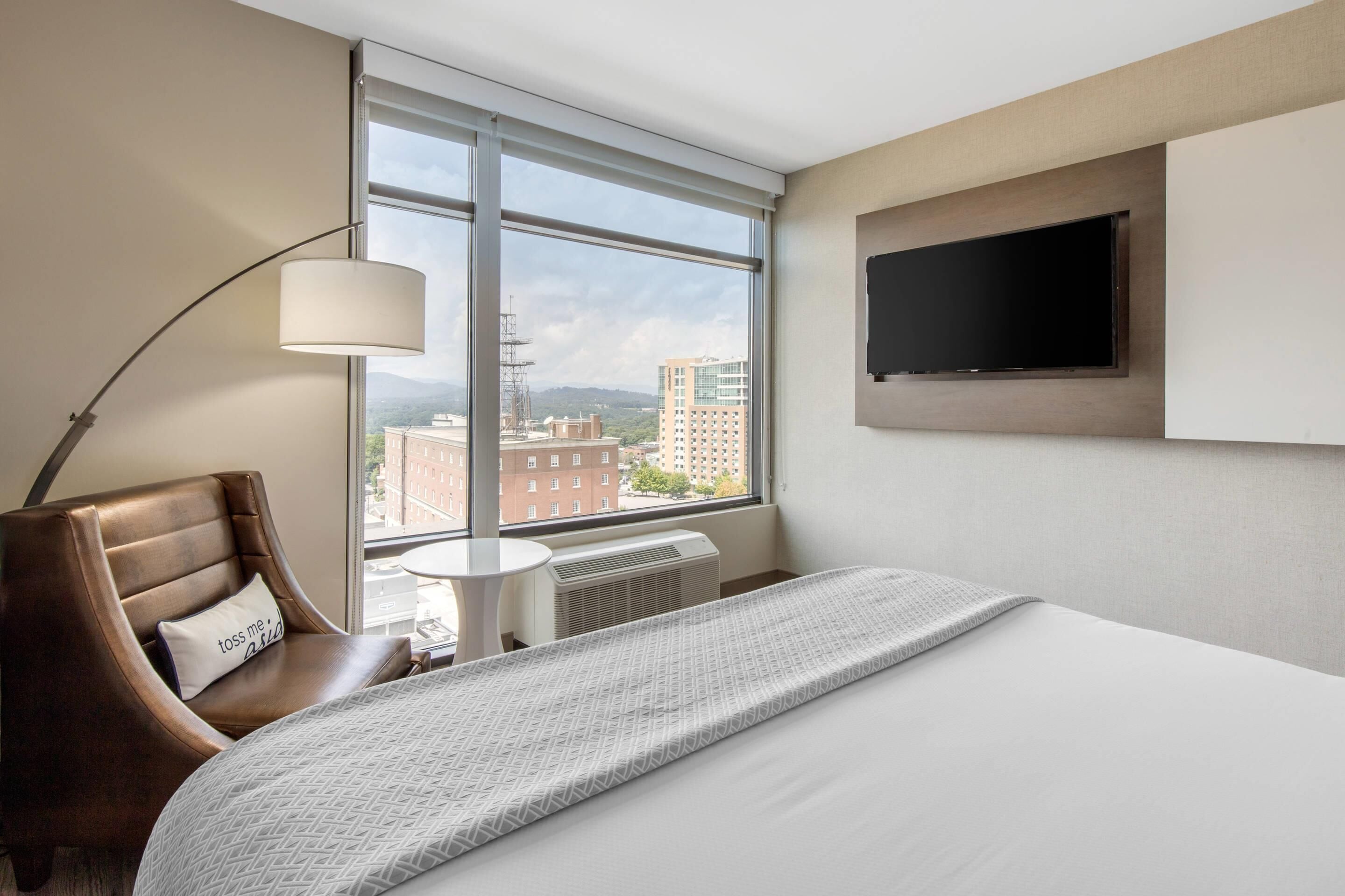 Habitación de hotel con cama, una butaca de cuero marrón, una mesita, una lámpara, televisor montado en la pared y ventana con vistas a edificios y montañas.