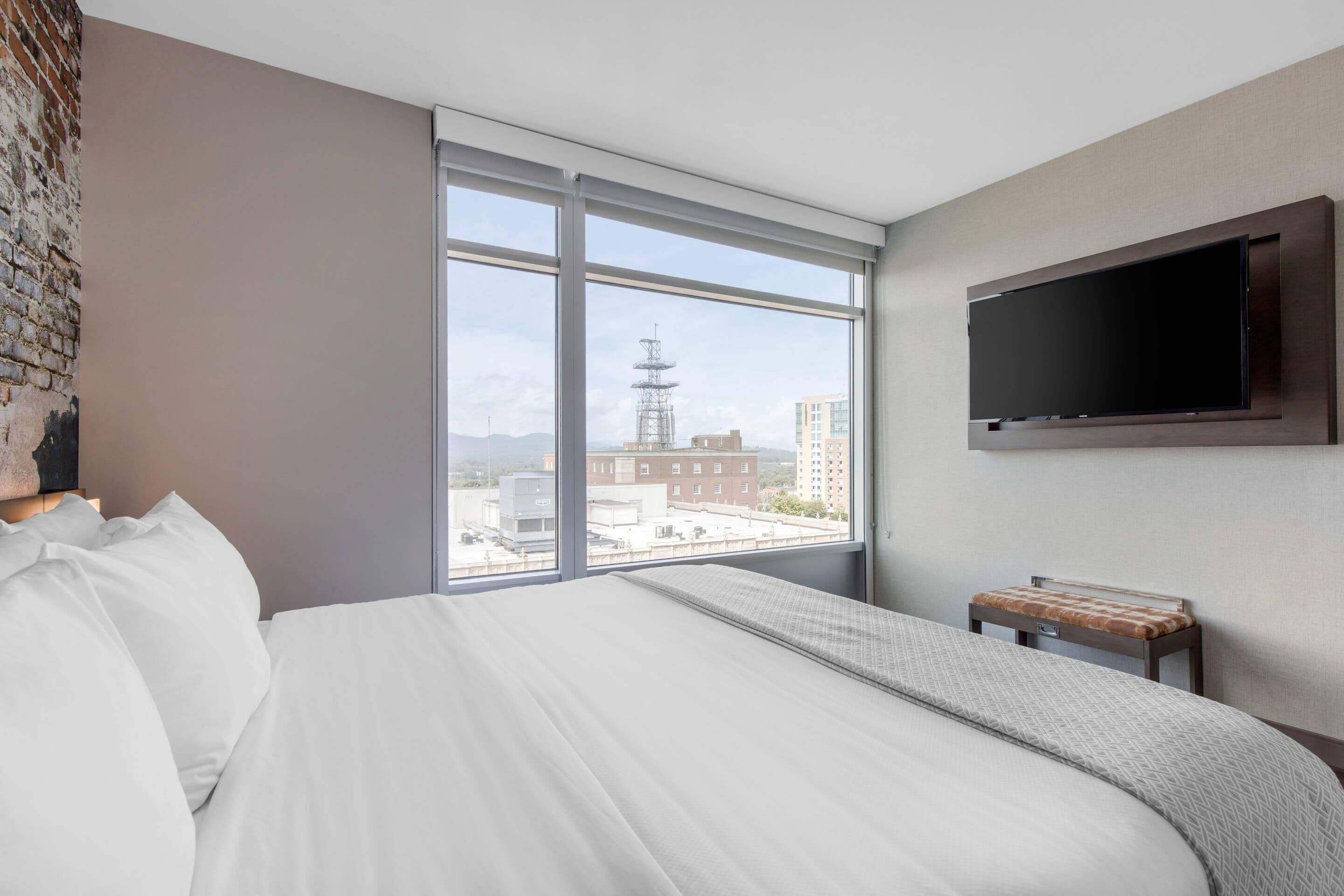Ett hotellrum med ett stort fönster, en säng, en tv som är monterad på väggen och ett litet bord med en kudde.