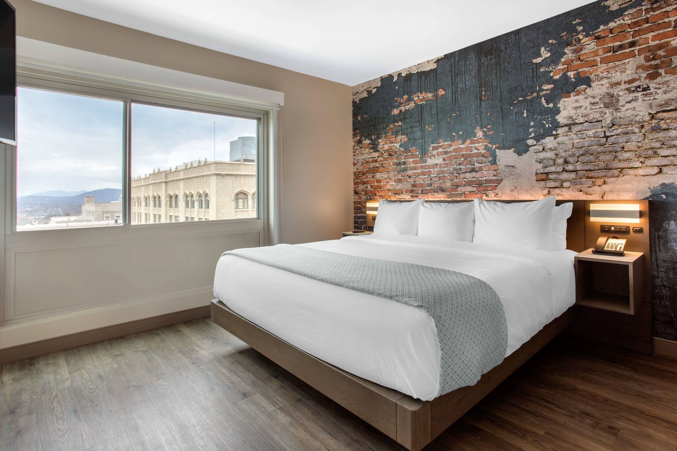 A modern hotel room with a large bed, a brick accent wall, a city view, and a wooden floor.