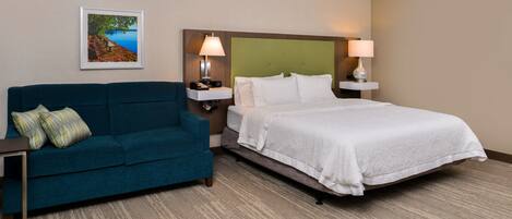 A hotel room with a bed, a blue sofa, a nightstand with a lamp, and a framed picture on the wall.