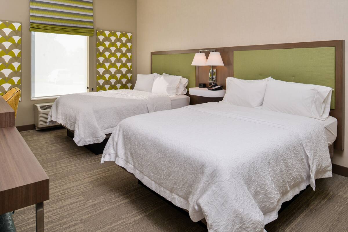 Habitación de hotel con dos camas, un escritorio, una ventana con persianas y una pared con diseño geométrico.