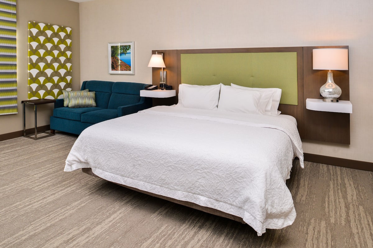 Habitación de hotel con una cama grande, un sofá azul, una mesita de noche con una lámpara y un estante de pared con otra lámpara.