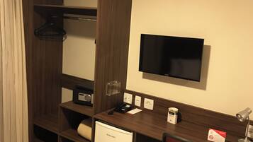 Habitación de hotel con escritorio de madera, un refrigerador pequeño, una silla, televisor montado en la pared y cama con cubrecama estampado.