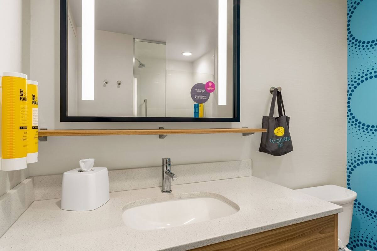 A bathroom with a mirror, sink, towel dispenser, and a towel rack with a bag.