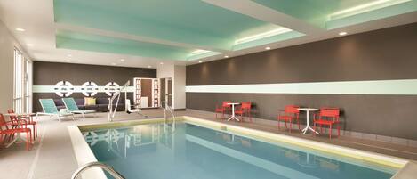 A swimming pool with a clear blue water, surrounded by red and blue chairs, and a ceiling with recessed lighting.
