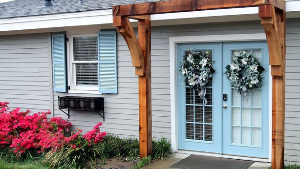 Una casa con puerta azul, un porche de madera y flores rosadas en el jardín.