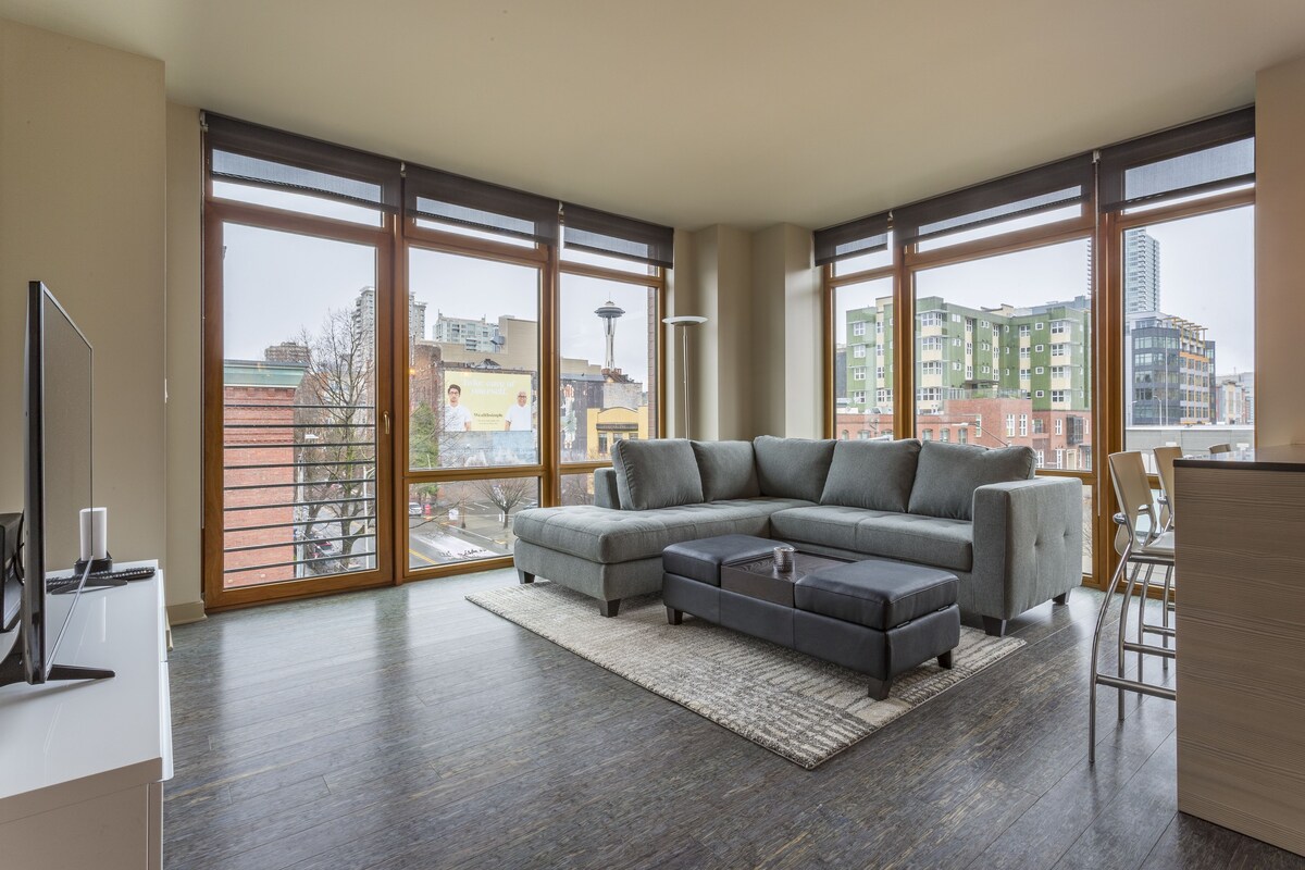 A modern living room with a sectional sofa, a flat-screen TV, and large windows offering a city view.