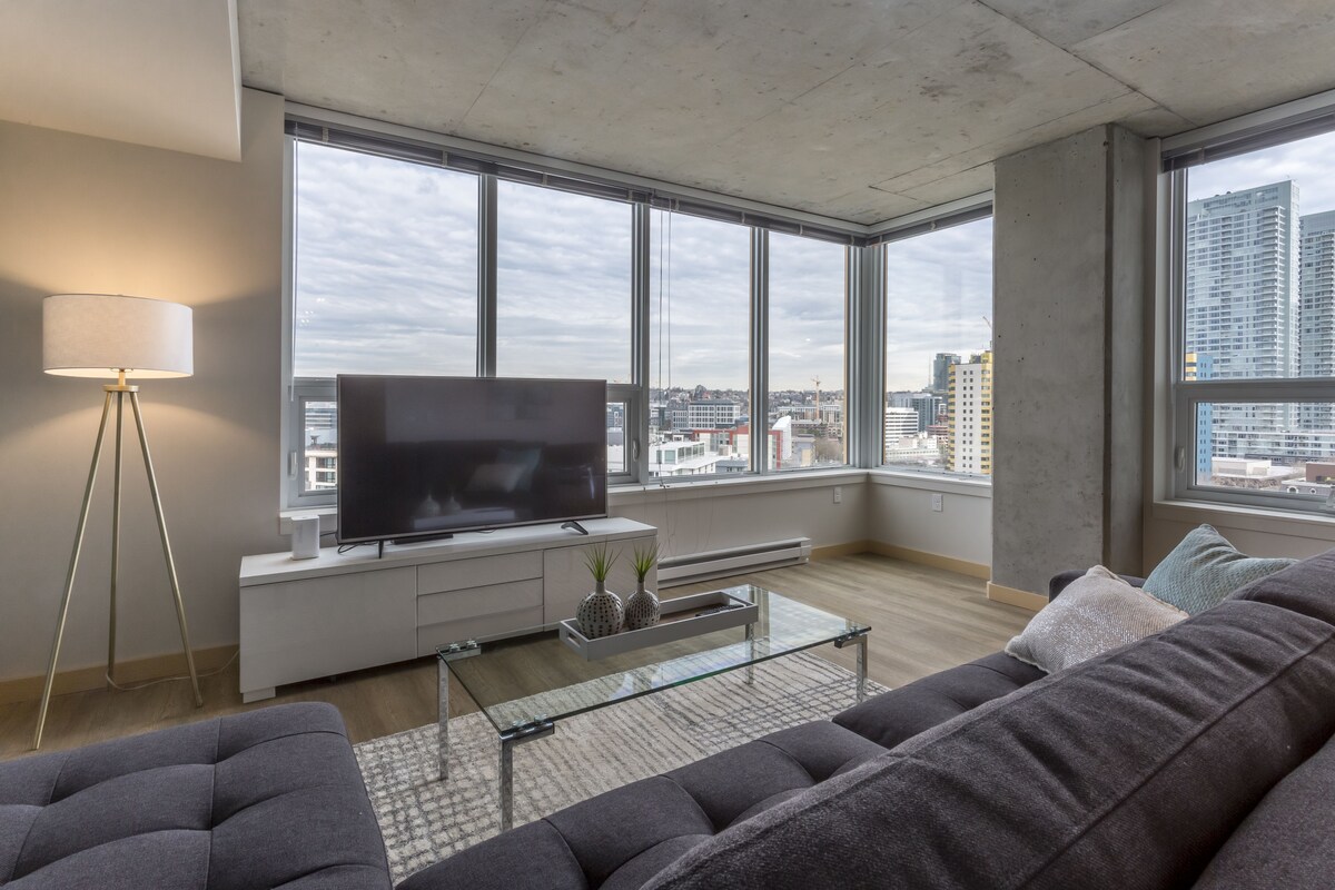 A modern living room with a large TV, comfortable seating, and large windows offering city views.