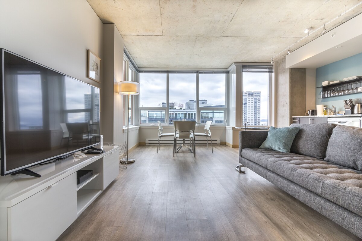 A modern living room with a large flat-screen TV, a dining area with chairs, a sofa, and a kitchen area with shelves.