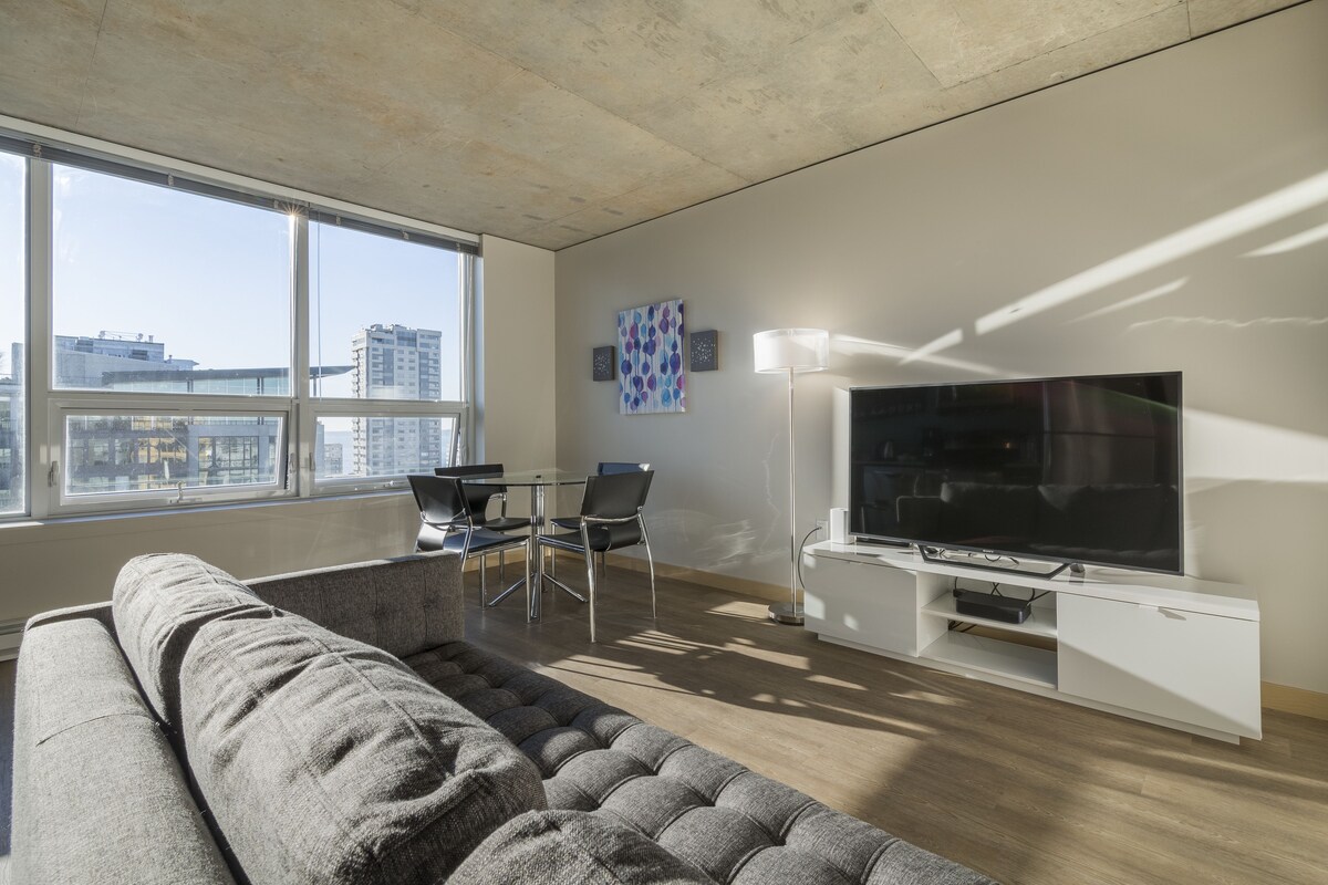 A modern living room with a large window, a grey sofa, a dining table with chairs, a flat-screen TV, and a white entertainment unit.