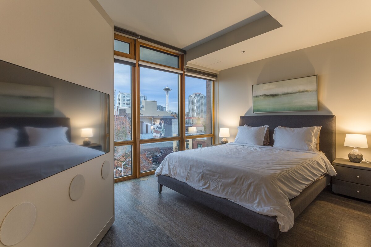 A modern hotel room with a large bed, a TV, and a city view through floor-to-ceiling windows.
