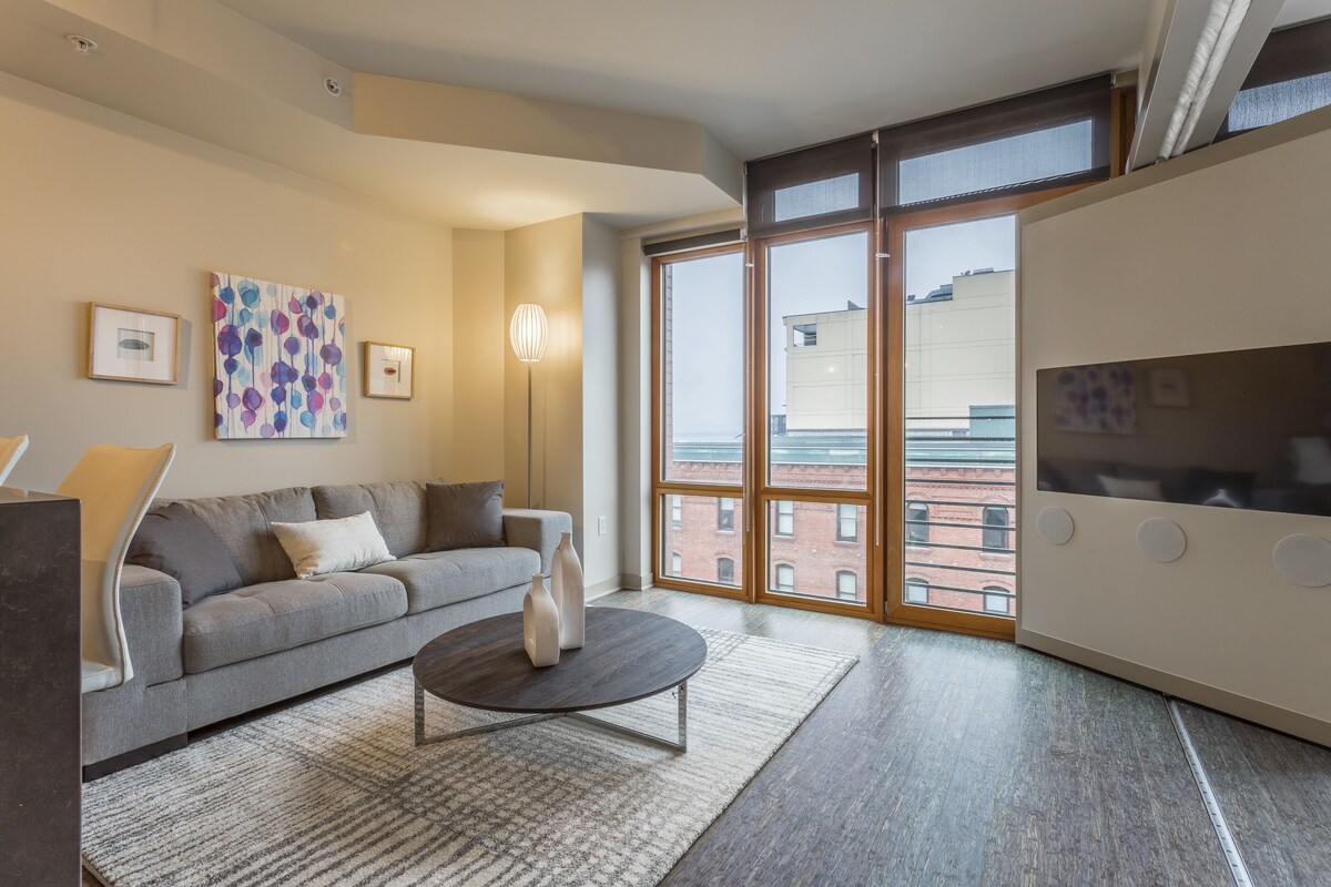 A modern living room with a grey sofa, a round coffee table, and a large window with a view of buildings.