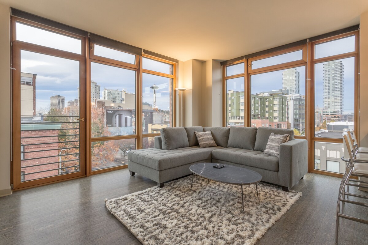 A modern living room with a sectional sofa, a coffee table, and a large window offering a city view.