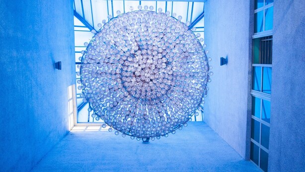 Escultura circular hecha con numerosos elementos pequeños en un pasillo con tonalidades azules y ventanas amplias.