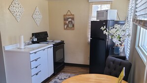 A compact kitchen with a white countertop, a black refrigerator, and a microwave. There's a dining table with a chair and a decorative wall hanging. - Lamplighter Cabins & Suites (Helena)