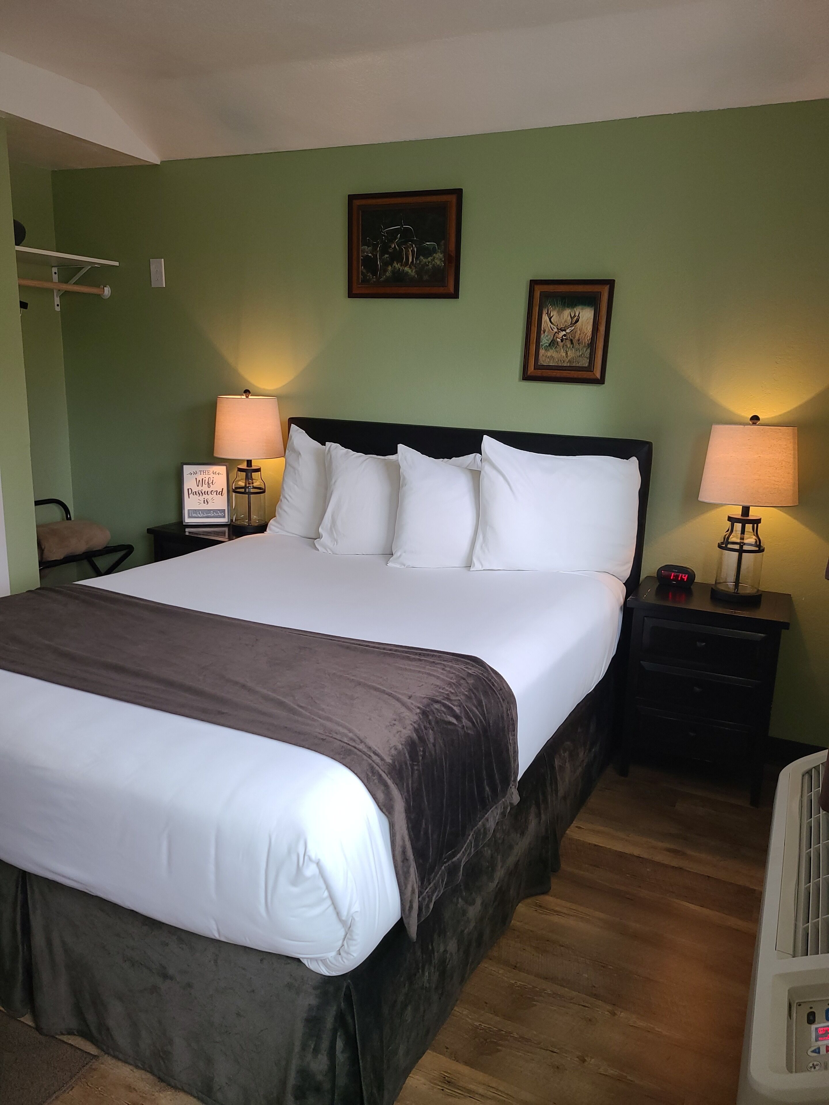 A neatly made bed with white linens and a brown bedspread, flanked by two bedside tables with lamps, in a room with green walls and wooden flooring.