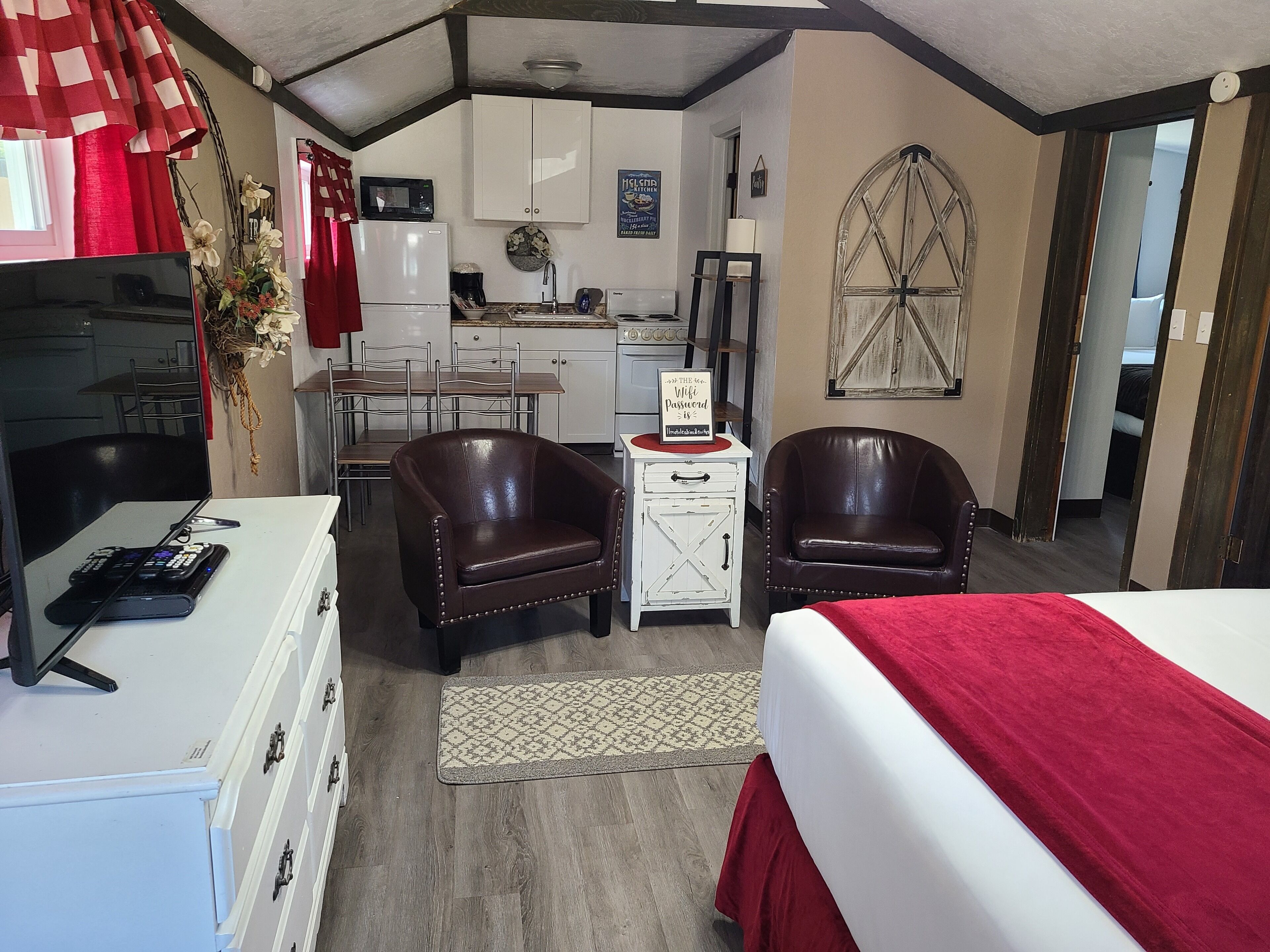 A compact room with a bed, two leather chairs, a small table, a desk, a microwave, a refrigerator, a sink, and a window with a decorative frame.
