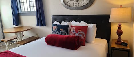 A hotel room with a bed, a round wall decoration, a window with curtains, a small round table, and a nightstand with a lamp.