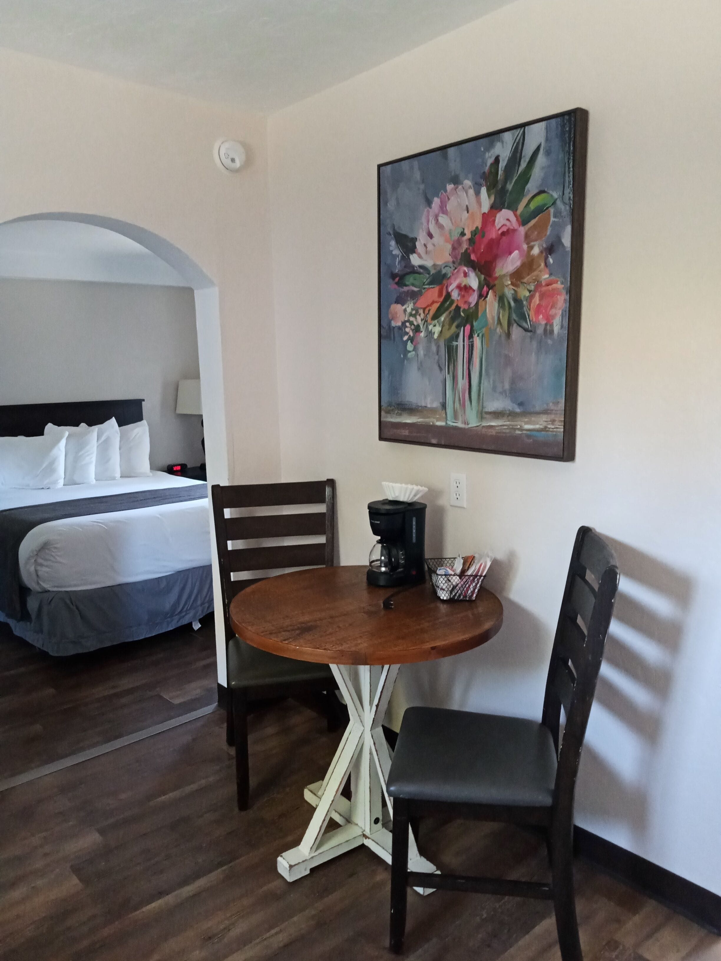 Habitación de hotel con una cama, una mesa de comedor con sillas, una cafetera y un cuadro de flores en la pared.