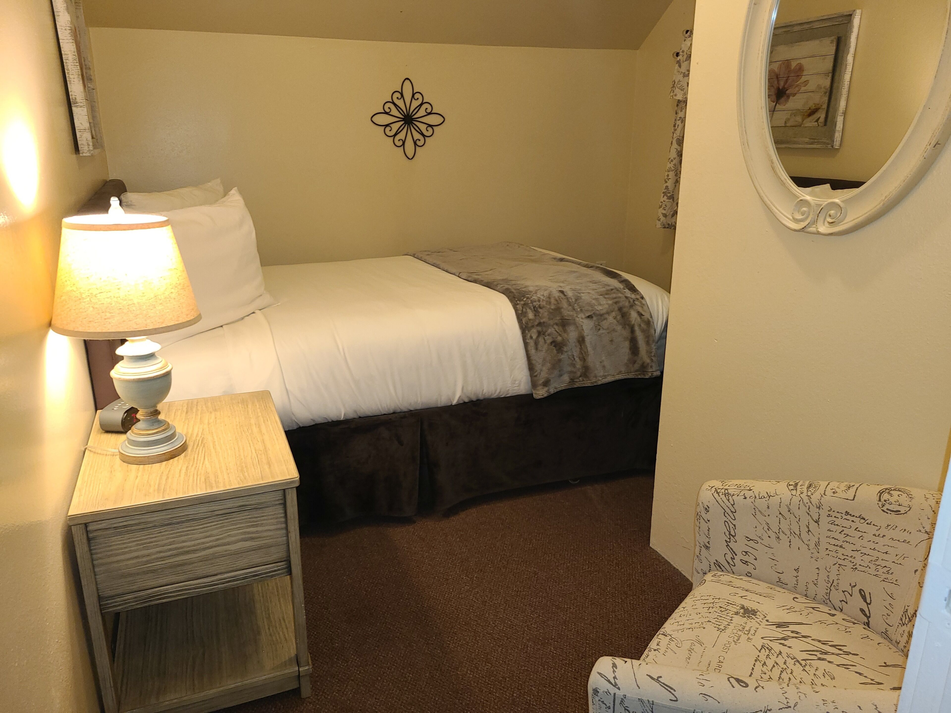 A bedroom with a bed, a bedside table with a lamp, a chair with a floral pattern, and a decorative wall piece.