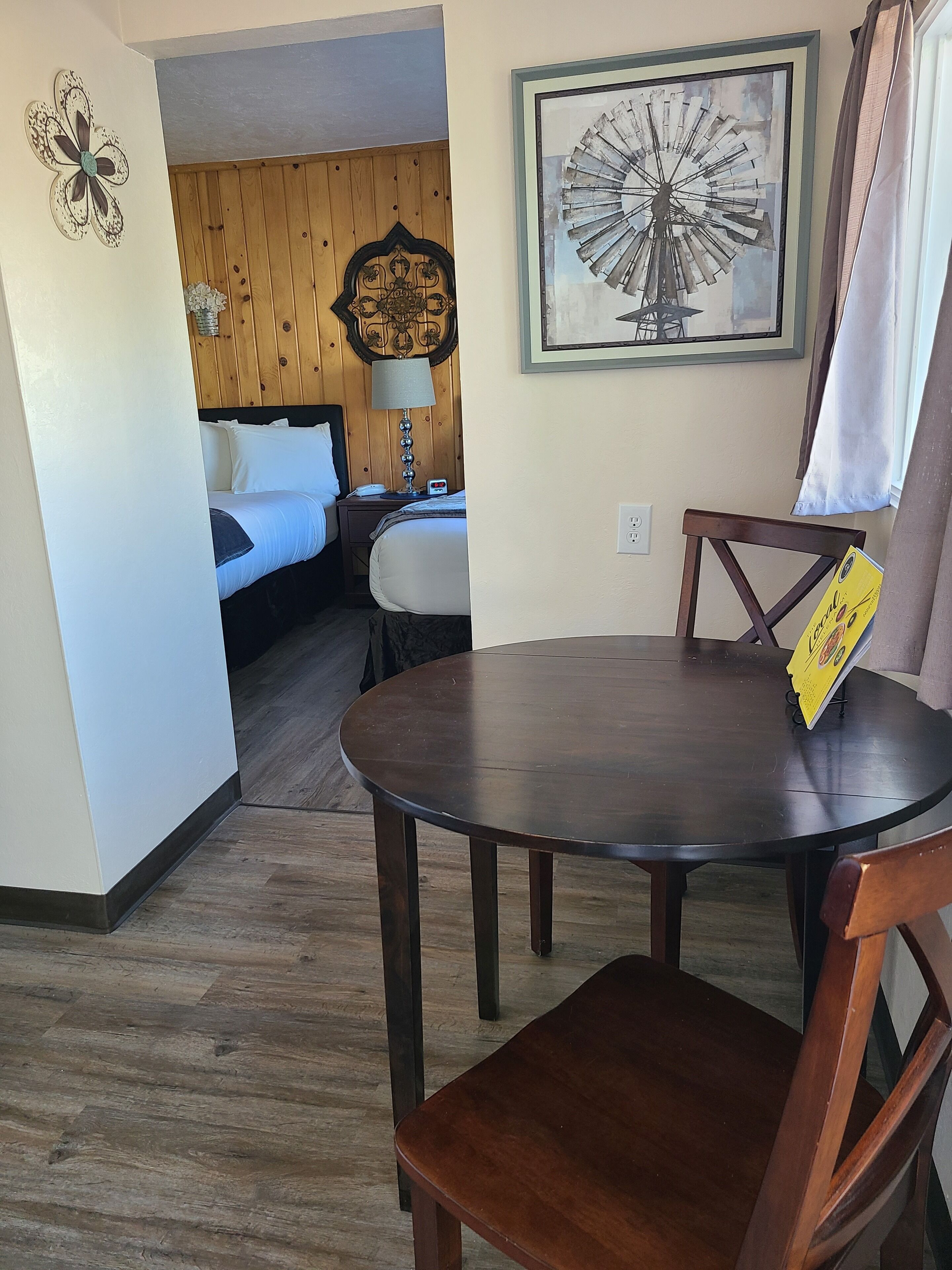 A hotel room with a wooden floor, a round dining table with chairs, a bed with white linens, a wooden headboard, a wall clock, a decorative wall piece, and a framed picture.