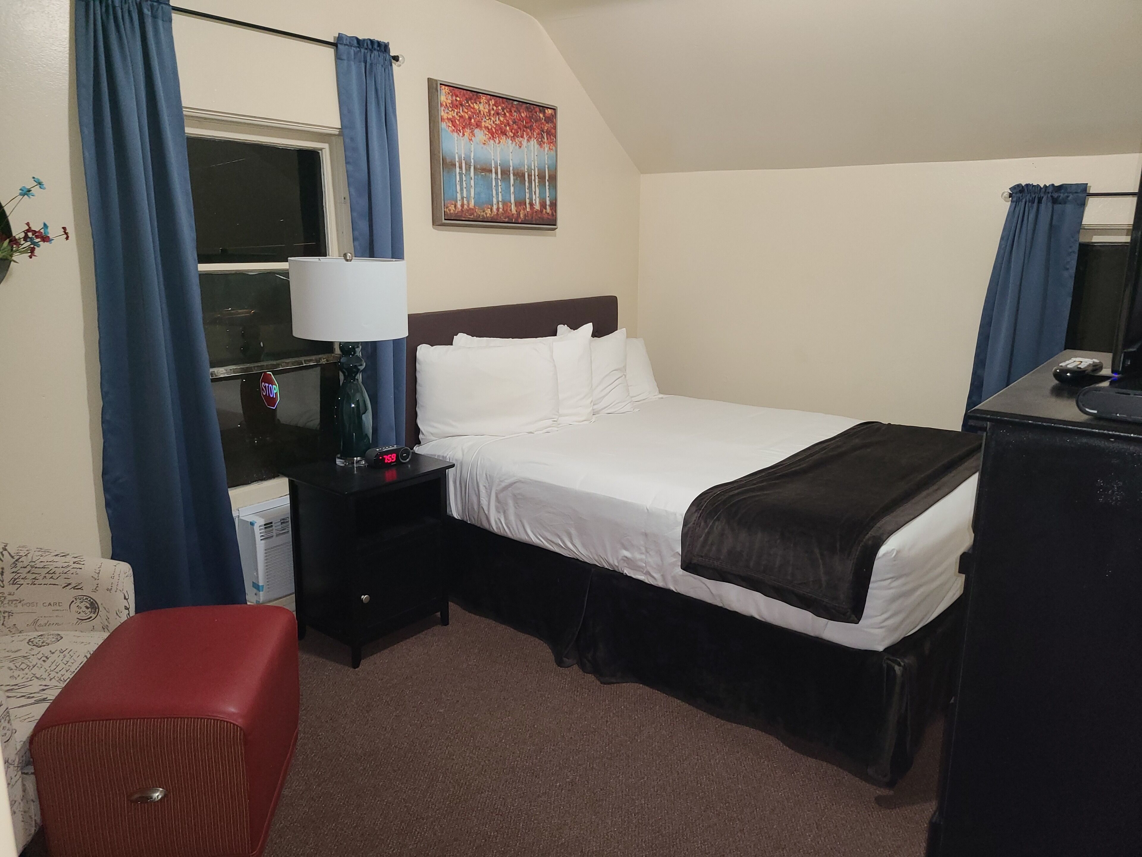 A hotel room with a bed, a nightstand, a lamp, a clock, a painting, and a desk with a computer.