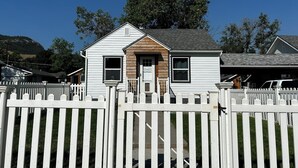 A small, single-story house with a white picket fence, a wooden front porch, and a mountain backdrop. - Lamplighter Cabins & Suites (Helena)