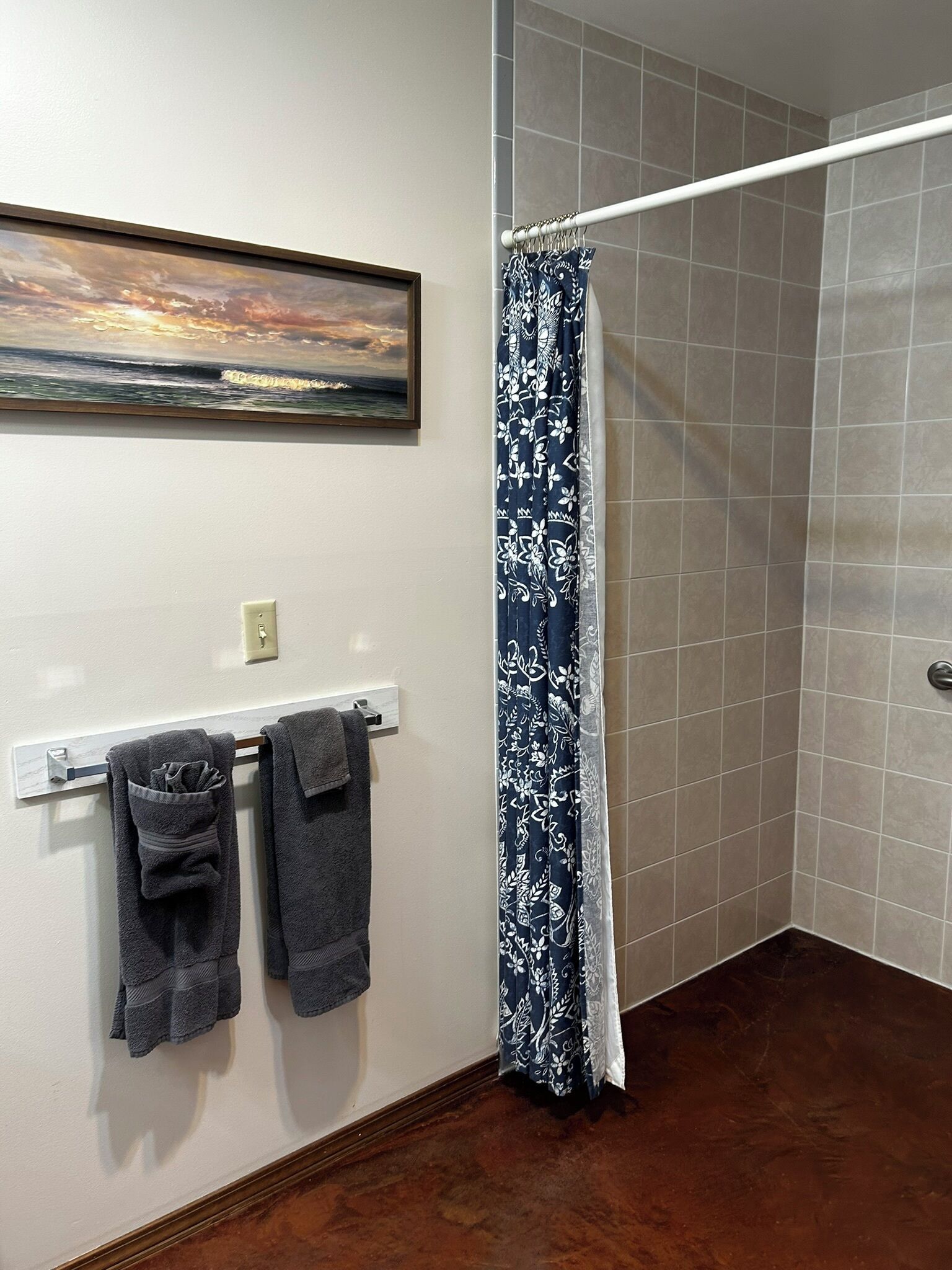 A bathroom with a shower, a towel rack with two towels, a painting on the wall, and a tiled floor.