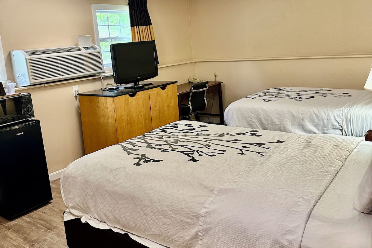 A hotel room with a bed, a desk with a computer, a mini-fridge, a heater, and a window with curtains.