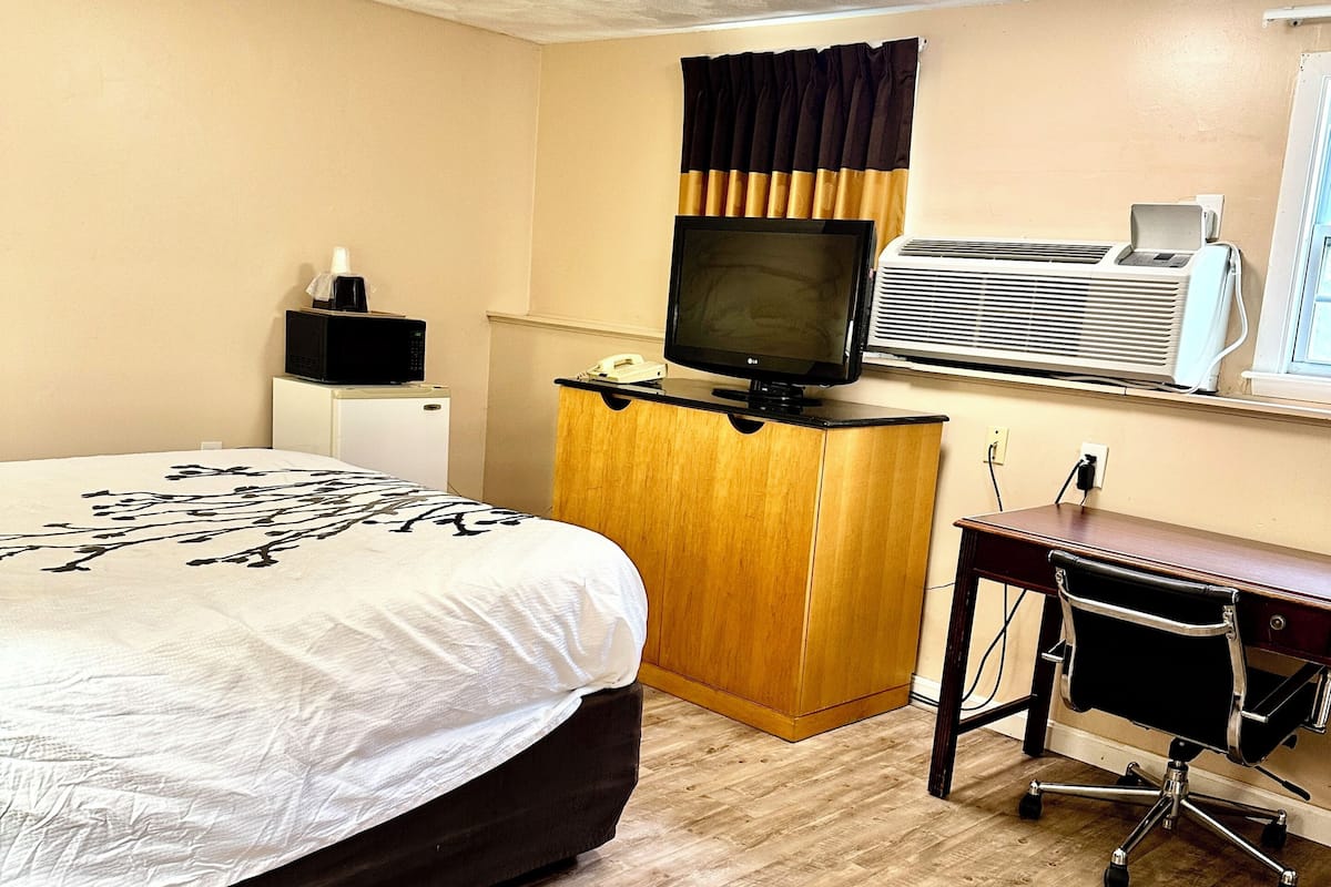 A hotel room with a bed, a desk with a computer, a television, and an air conditioning unit.