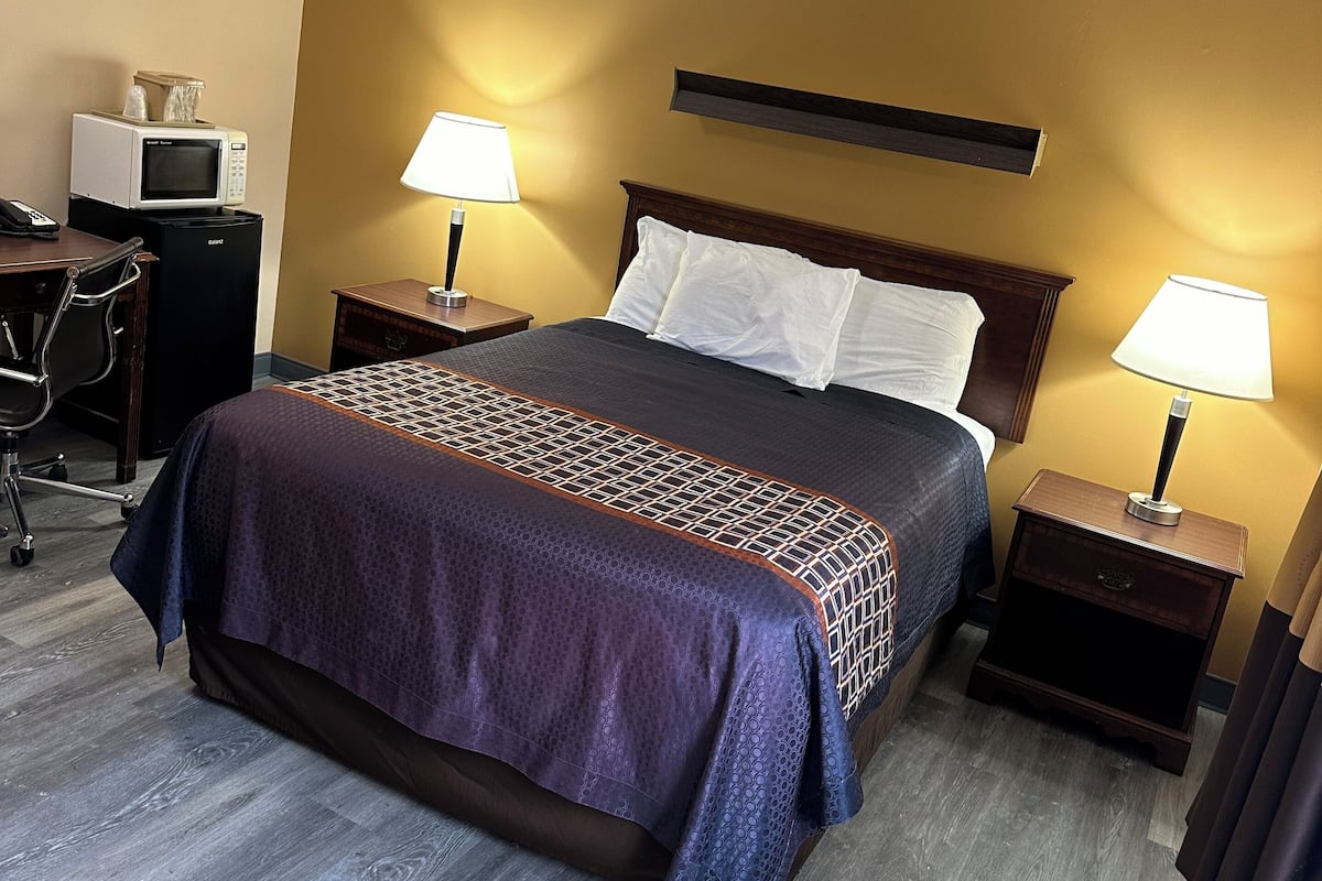 A hotel room with a bed, two bedside tables with lamps, a microwave, a refrigerator, and a desk with a chair.
