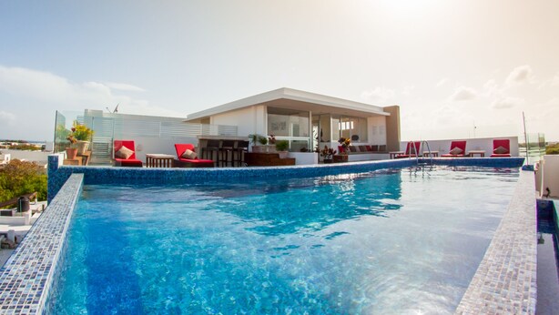 A rooftop infinity pool with red lounge chairs and a modern building with large windows. - In heart of town 1BR condo in steps distance from the 5th Av by Happy Address (Playa del Carmen)