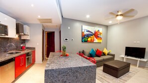 A modern kitchen with granite countertops, red cabinetry, and a central island. There is a dining area with a sofa and a flat-screen TV. - In heart of town 1BR condo in steps distance from the 5th Av by Happy Address (Playa del Carmen)