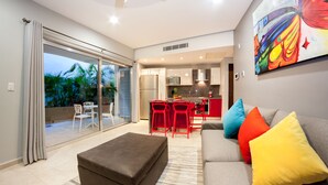 A modern living room with a grey sofa, colorful cushions, a brown ottoman, and a glass door leading to a balcony with a view of palm trees. - In heart of town 1BR condo in steps distance from the 5th Av by Happy Address (Playa del Carmen)