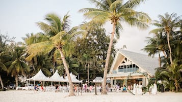 A beachside venue with white tents, palm trees, and a building with a thatched roof.