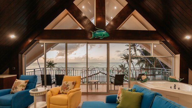 A spacious living area with a large window overlooking a balcony with a view of the sea. There are blue and yellow sofas, a small round table, and a large bathtub. - Surfrider Resort Hotel (Saipan)