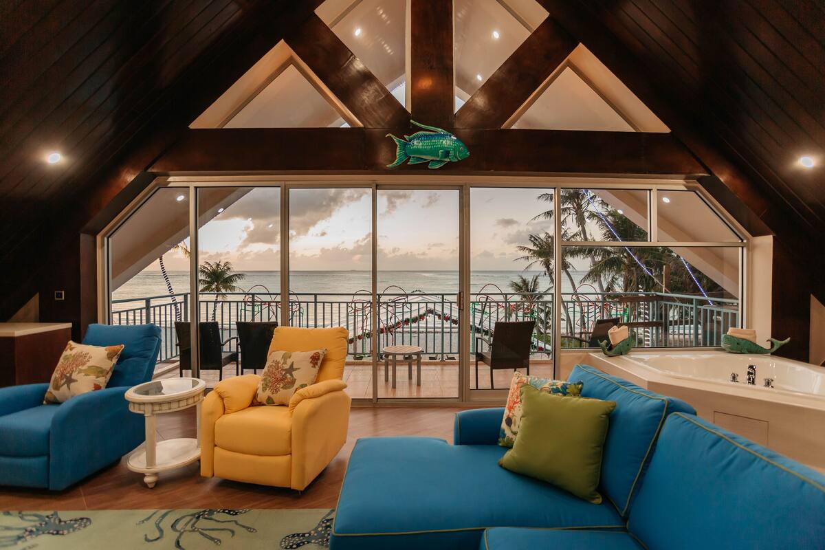 A spacious living area with a large window overlooking a balcony with a view of the sea. There are blue and yellow sofas, a small round table, and a large bathtub.