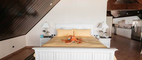 A bedroom with a white bed, a wooden ceiling, and a view into the kitchen.