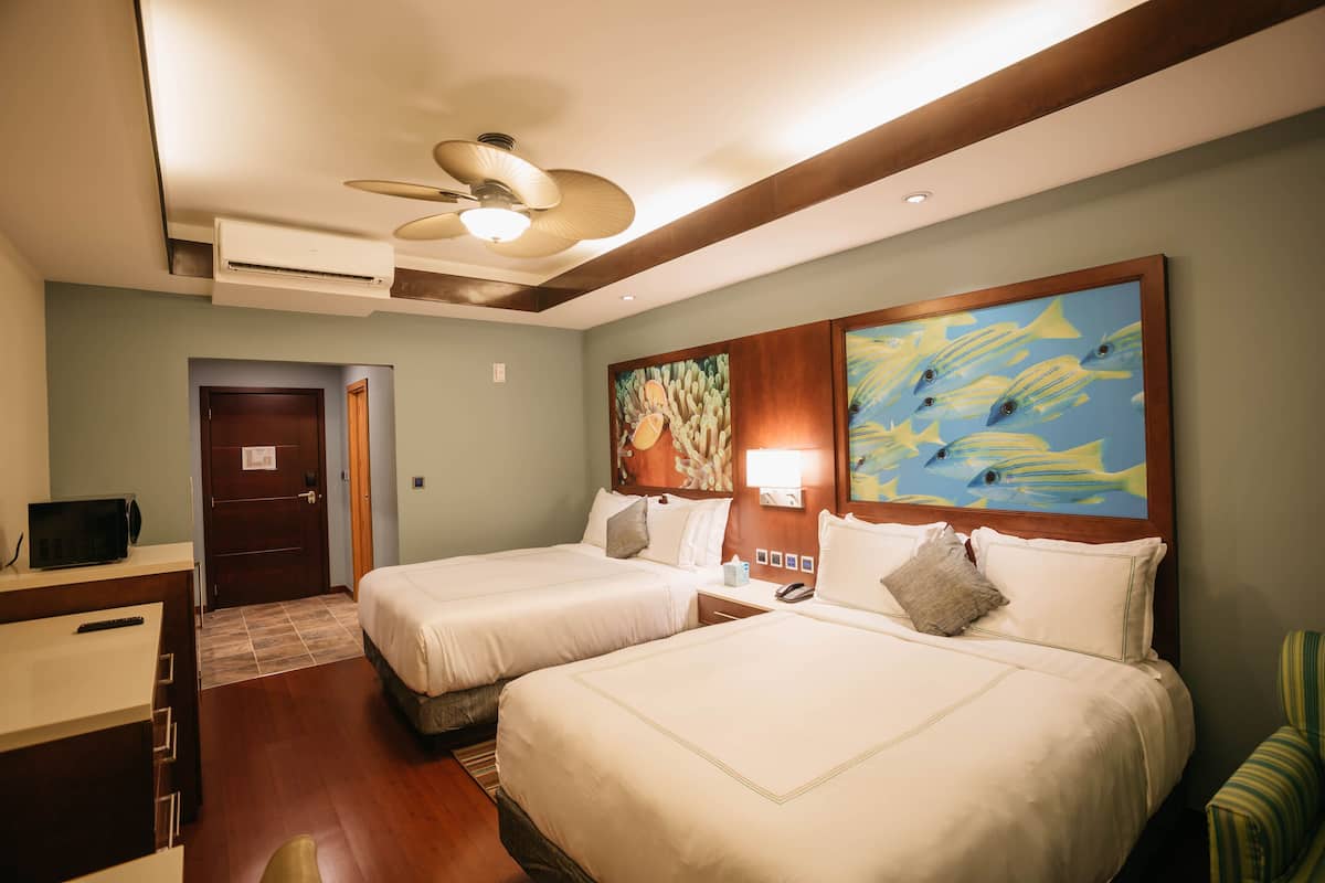 A hotel room with two beds, a desk with a computer, a ceiling fan, and artwork on the walls.