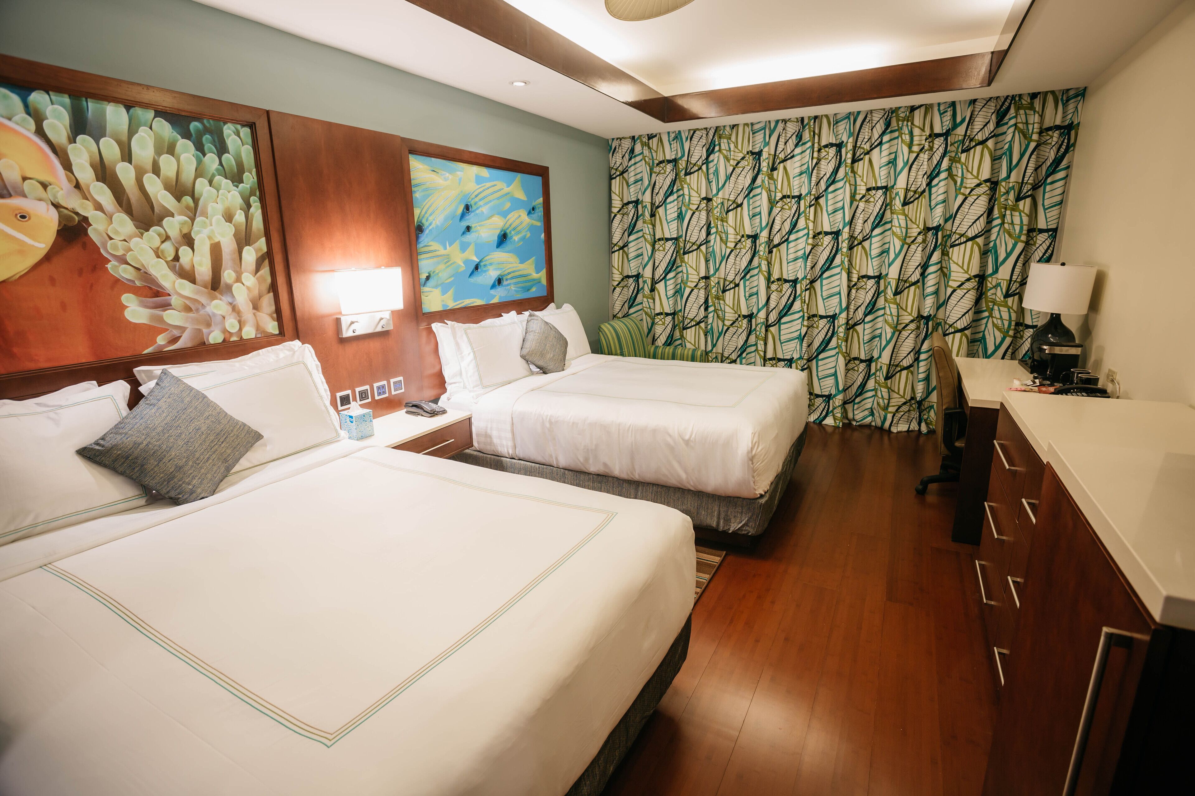 A hotel room with two beds, a wooden headboard, a painting of fish, a patterned curtain, and a desk with a lamp.